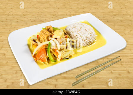 Chinesisches Essen auf einem weißen Teller auf einem Tisch stehend. Gebratenes Schweinefleisch mit Reis, Gemüse und Kokos-Sauce. Hintergrund-Holz. Stockfoto