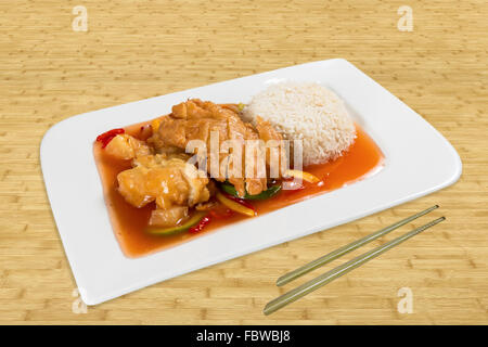 Chinesisches Essen auf einem weißen Teller. Gebratenes Huhn mit Gemüse, Reis und roter Soße. Hintergrund Holz mit Stäbchen Stockfoto