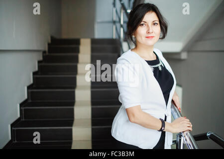 Erfolgreiche attraktive Brünette mit freundlichen Augen stehend auf der Treppe des Bürogebäudes, während einer Pause bei der Arbeit. Weiße Jacke und strengen schwarzen Kleid Schmuck tragen. Nette junge Frau posiert. Stockfoto