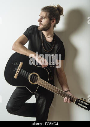 Foto von einem jungen attraktiven Mann mit langen Haaren und Bart lehnte sich gegen die Wand mit einer akustischen Gitarre. Stockfoto