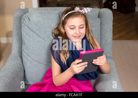 Junges Mädchen im rosa Kleid sitzt auf einem Stuhl, ein e-Book-Reader oder Tablet ist farblich mit ihrem Outfit zu lesen. Stockfoto