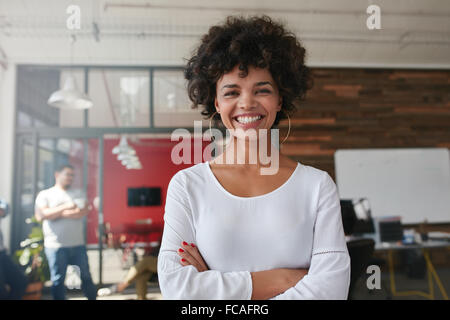 Junge Frau, die Arme gekreuzt und Blick in die Kamera Lächeln. Sie steht in einem modernen Büro mit ihren Kollegen Stockfoto