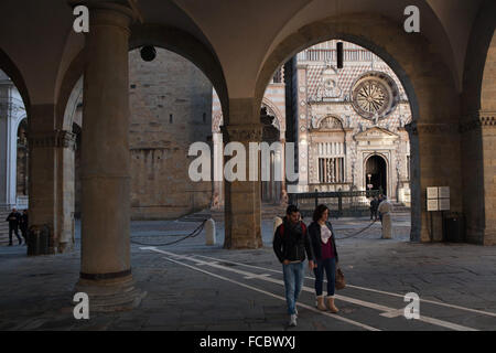 Die Menschen gehen durch die Arkade der Palazzo della Ragione am Piazza Vecchia in Bergamo, Lombardei, Italien, mit der Kirche Santa Maria Maggiore (L) und die Cappella Colleoni (R) im Hintergrund zu sehen. Stockfoto