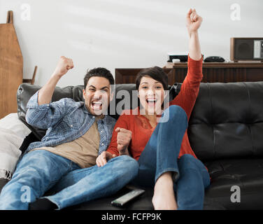 Mann und Frau auf einem Sofa sitzen, feiern und Pumpen die Luft mit ihren Fäusten. Sport auf dem Fernseher ansehen. Stockfoto