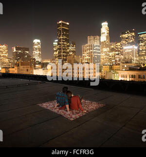 Ein paar sitzt auf einem Teppich auf eine Dachterrasse mit Blick auf eine Stadt beleuchtet in der Nacht. Stockfoto