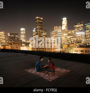 Ein paar sitzt auf einem Teppich auf eine Dachterrasse mit Blick auf eine Stadt beleuchtet in der Nacht. Stockfoto