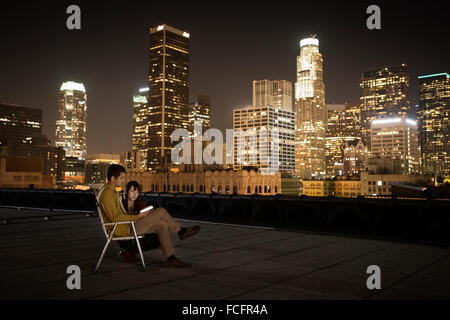 Ein paar auf einer Dachterrasse mit Blick auf Los Angeles bei Nacht, man sitzt, ein Smartphone zu betrachten. Stockfoto