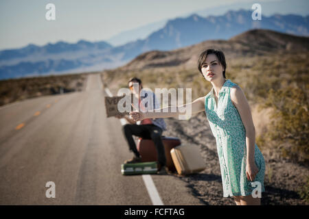 Ein junges Paar, Mann und Frau auf eine asphaltierte Straße in der Wüste Hitchiking, mit einem Schild mit der Aufschrift Vegas oder pleite. Stockfoto