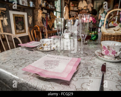 Rosa Bridal Dusche Serviette Gedeck mit Champagne Flute und Tee Tassen auf Spitzen Tischdecke in einem Raum voller Nippes. Stockfoto