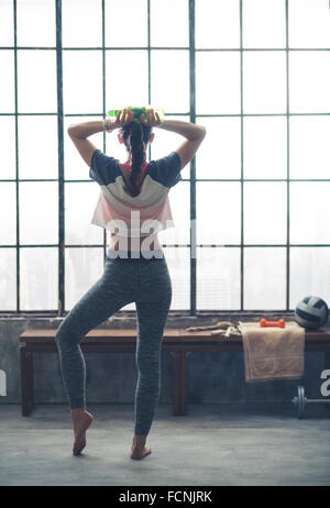 Von hinten gesehen, steht eine Frau, einen Fuß lässig ausstrecken, wie sie eine Flasche Wasser, hält ihre Hände hinter ihrem Kopf ruht. Stockfoto