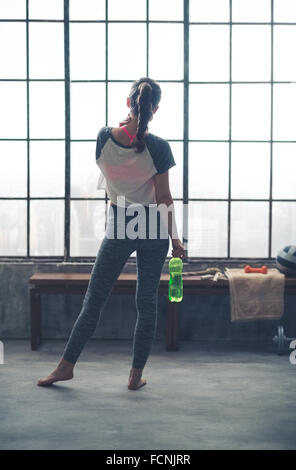 Halten eine Flasche Wasser an ihrer Seite, eine Passform steht sportliche Frau, schaut aus dem Loft Fitness-Studio-Fenster in der Stadt unten. Barfuß, Stockfoto
