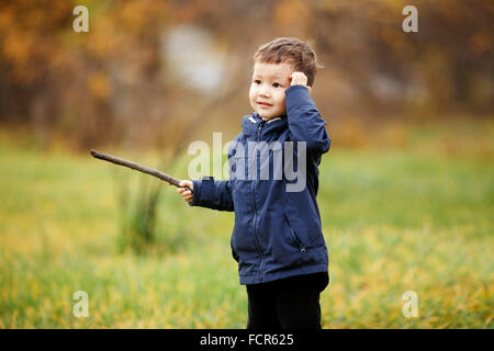 Netter Junge mit Holzstab in der Hand spielen im Herbst Park im Freien. Verwirrt, denken, was zu tun ist, auf der Suche zur Seite. Herbst, gelbe Blätter. Kinder tragen blaue Jacke. Unbeschwerte Kindheit. Stockfoto