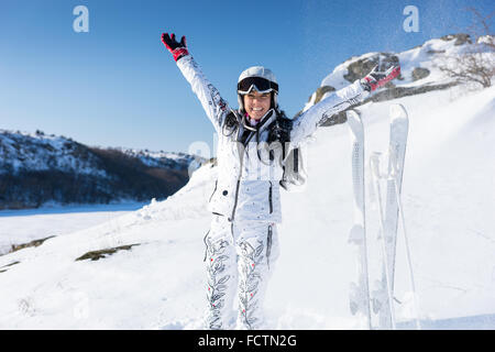 Begeisterte junge Erwachsene Skifahrer in lange Haare und weißen Schneeanzug Schnee über ihrem Kopf mit aufrechten Skiern auf Hügel hinter ihr zu werfen Stockfoto