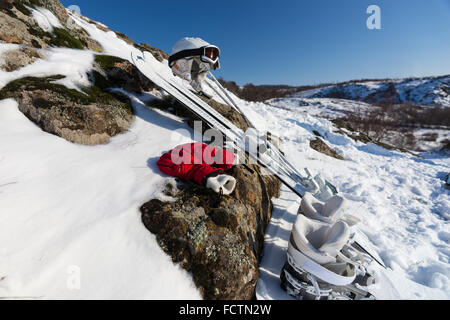 Saubere Darstellung der weißen Stiefeln, Paar Ski, Helm und rote Handschuhe platziert auf dem Schnee bedeckt Boulder unter blauem Himmel auf sonnigen Wintertag. Stockfoto