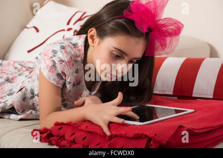 Kleine Mädchen spielen mit einem Tablet-PC im Wohnzimmer zu Hause, wie sie auf eine bunte rote Decke auf dem Sofa Surfen liegt Stockfoto