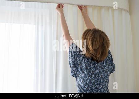 Frau, die seine weiße Vorhänge am Fenster aufhängen Stockfoto