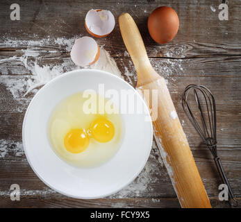 Kochen oder backen. Stillleben mit Eiern, rohen Eiern in eine Schüssel, Eierschalen, Mehl, Nudelholz und Schneebesen auf hölzernen Hintergrund. Stockfoto