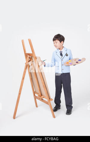 Elementary School Boy in Schuluniformen stehend und zeichnen mit einem Pinsel und einer Palette auf ein Sketch Pad auf einer Staffelei Stockfoto