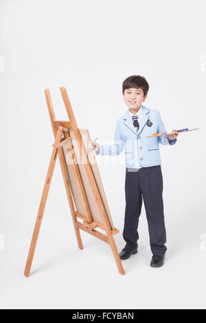 Elementary School Boy in Schuluniformen stehend und zeichnen mit einem Pinsel und einer Palette auf ein Sketch Pad auf einer Staffelei Stockfoto