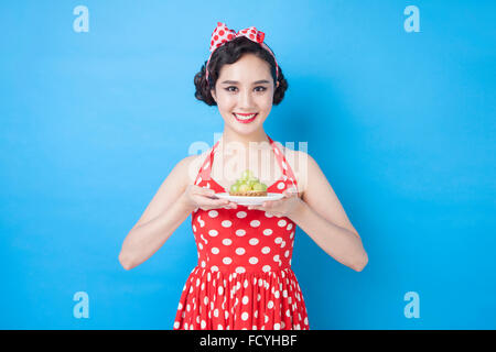 Frau im Retro-Stil Mode hält eine Frucht Schale Teller in ihren Händen und starrte nach vorne mit einem Lächeln auf blauem Hintergrund Stockfoto
