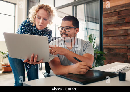 Porträt von junger Mann sitzt an seinem Schreibtisch zeigt auf Laptop in Händen von einer Kollegin, männliche Grafiker helfen ein Weibchen Stockfoto