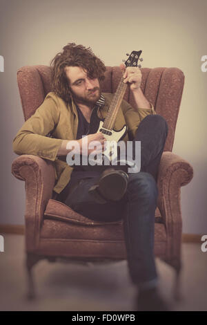 Bärtige Musiker spielt elektrische Ukulele Gitarre in Vintage Sessel Stockfoto