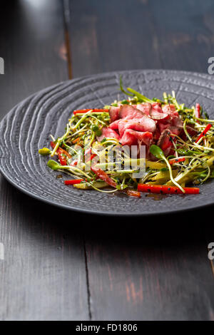 Authentische Salat mit Speck, frische rote Paprika, Sprossen, Sesam und Olivenöl auf einer schwarzen Platte. Morgen stimmungsvolle Beleuchtung, Stockfoto