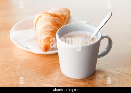 Cappuccino mit Croissant. Tasse Kaffee mit Milchschaum steht auf Holztisch, Nahaufnahme Foto mit selektiven Fokus Stockfoto