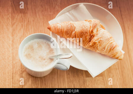 Cappuccino mit Croissant. Tasse Kaffee steht auf Holztisch in Kantine, Nahaufnahme Foto mit selektiven Fokus Stockfoto