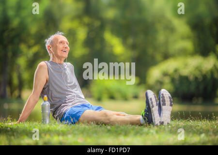 Aktive senior woman Sportswear im Park sitzen und hören von Musik über Kopfhörer mit Tilt-Shift-Objektiv geschossen Stockfoto