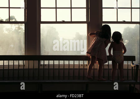 Zwei junge Mädchen auf einer langen Bank schaut aus einem nebligen Fenster mit dem Rücken zur Kamera stehen. Stockfoto