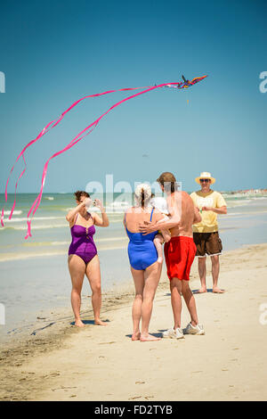 Frau nimmt Foto des Paares mit Baby am Strand, während ein Mann einen overhead rosa Drachen mit Streamer in der Nähe der Brandung fliegt Stockfoto