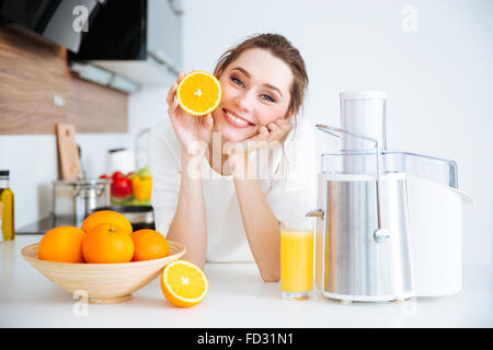 Fröhliche hübsche junge Frau zeigt die Hälfte der Orange in der Küche sitzen Stockfoto