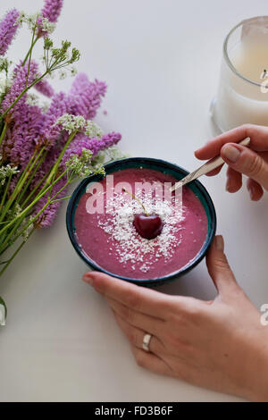 Pink Cherry und Kokos Smoothie in eine Schüssel geben, garniert mit Kokosraspeln und frische Kirschen, lila und weißen Blumen auf dem Tisch Stockfoto