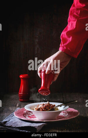 Weibliche Hand wirft Salz aus roten Salzstreuer in Vintage englische Teller hausgemachter Pasta Pici mit Bolognese Sauce, serviert auf g Stockfoto