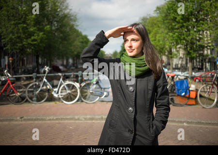 Schöne junge Frau, bewundern Sie die Schönheit von Amsterdam auf einer Brücke schützen ihre Augen vor Sonne mit Hand gekleidet im schwarzen Mantel ein Stockfoto