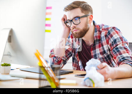 Gelangweilt erschöpft bärtiger junger Mann im karierten Hemd sitzen am Arbeitsplatz und Blick auf monitor Stockfoto
