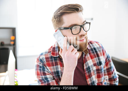 Glücklich attraktiven jungen Mann mit Bart im karierten Hemd und Brille reden über Handy im Büro