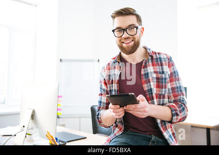 Fröhliche attraktiver junger Mann mit Bart im karierten Hemd und Brille sitzt und hält Tablet im Büro Stockfoto