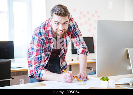 Attraktives Lächeln bärtiger Mann im karierten Hemd stehend und auf Blaupause im Büro arbeiten Stockfoto