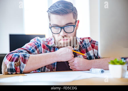 Porträt von nachdenklich bärtiger junger Ingenieur in Gläser am Tisch sitzen und Plan Stockfoto