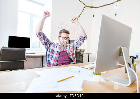 Erfolgreiche glücklich in Gläsern mit erhobenen Händen sitzen am Arbeitsplatz im Büro Stockfoto