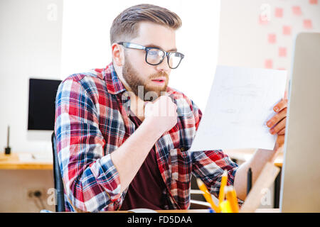 Ernst gut aussehender Mann mit Bart in Gläsern sitzen und mit Blick auf Blaupause im Büro Stockfoto