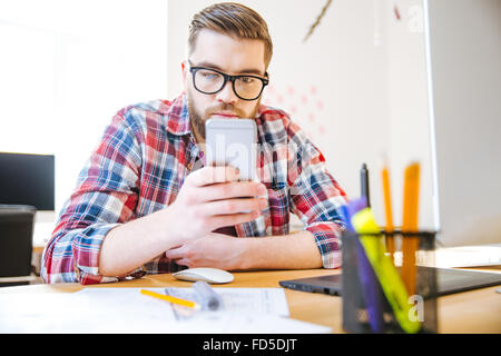 Hübscher konzentrierte junger Mann mit Bart im karierten Hemd am Tisch sitzen und mit Handy Stockfoto