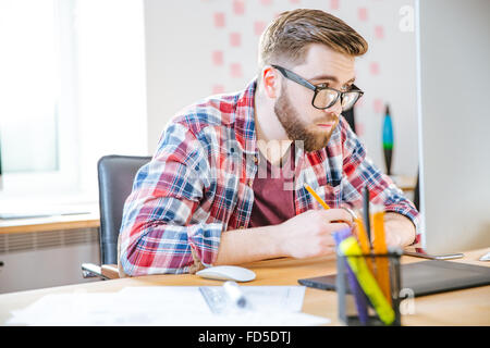 Ernst gut aussehender Mann mit Bart im karierten Hemd mit Tablet-PC am Arbeitsplatz sitzen und mit Blick auf Gläser Stockfoto