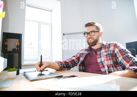 Lächeln auf den Lippen schön bärtigen Jungdesigner im karierten Hemd und Brille, Zeichnung und Grafik Tablett und Stift Stockfoto