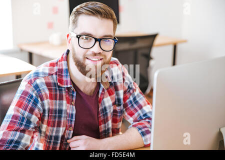 Glücklichen hübscher junger Mann mit Perle im karierten Hemd und Brille sitzen am Arbeitsplatz und die Arbeit mit computer Stockfoto