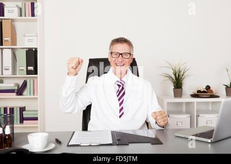 Jubelnde mittleren Alters männlichen Arzt sitzen Jubel in seinem Büro Stanzen der Luft mit der Faust, wie er einen Erfolg feiert Stockfoto