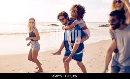 Zwei glückliche junge Männer geben ihre Freundinnen Huckepack tragen am Strand. Gruppe von Freunden Urlaub am Meer zu genießen. Stockfoto