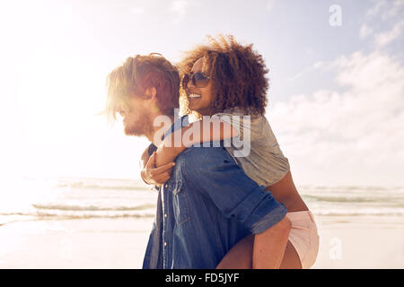 Seite Ansicht Porträt junger Mann mit seiner Freundin auf dem Rücken am Strand. Freunde geben Huckepack Fahrt zu seinen schönsten Stockfoto